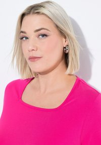 Camiseta fucsia con cuello redondo, hecha de tela suave. Combinada con pendientes metálicos grandes, cabello corto rubio y maquillaje neutro.