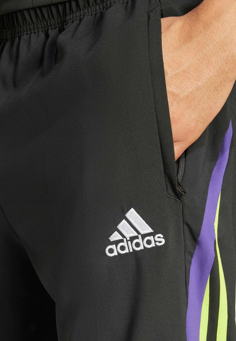 Pantalones deportivos negros con un acabado texturizado, que presentan un logo de Adidas en blanco y rayas verticales en púrpura y verde a los lados.