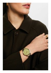 Goldfarbener Uhr mit grünem Ziffernblatt, silbernen Stundenmarkierungen und einem Metallarmband. Trägt sie an einer Hand, die auf einem dunklen Stoff ruht.