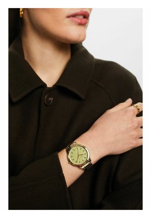 Goldfarbener Uhr mit grünem Ziffernblatt, silbernen Stundenmarkierungen und einem Metallarmband. Trägt sie an einer Hand, die auf einem dunklen Stoff ruht.