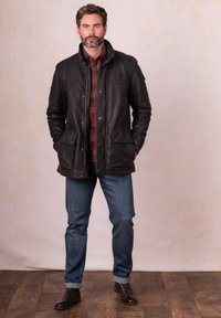 Brown leather jacket with a collar, two front pockets, and snap buttons, layered over a red and plaid shirt, worn with blue jeans and brown boots.