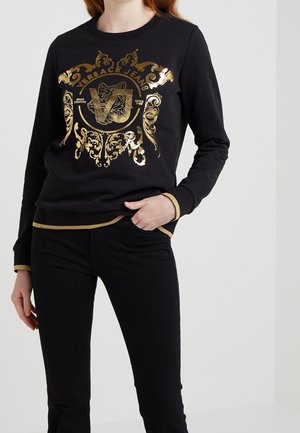 Person iført sort Versace Jeans sweatshirt med gyldent udsmykket våbenskjold og sorte bukser, hånd hvilende i forlomme.