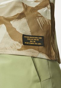 Kamikaze-paita beige- ja vaaleanruskeassa sävyssä mustalla etiketillä, joka näyttää tuote tiedot; yhdistettynä vaaleanvihreisiin kangashortsseihin, joissa on tasku.