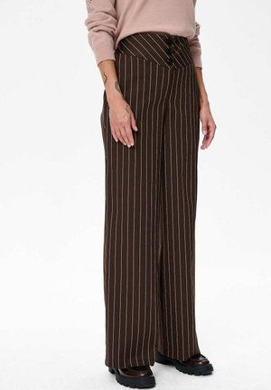 Frau trägt braune, weit geschnittene, hoch taillierte Hose mit weißen Streifen, drei schwarzen Knöpfen am Bund und schwarzen klobigen Schuhen.