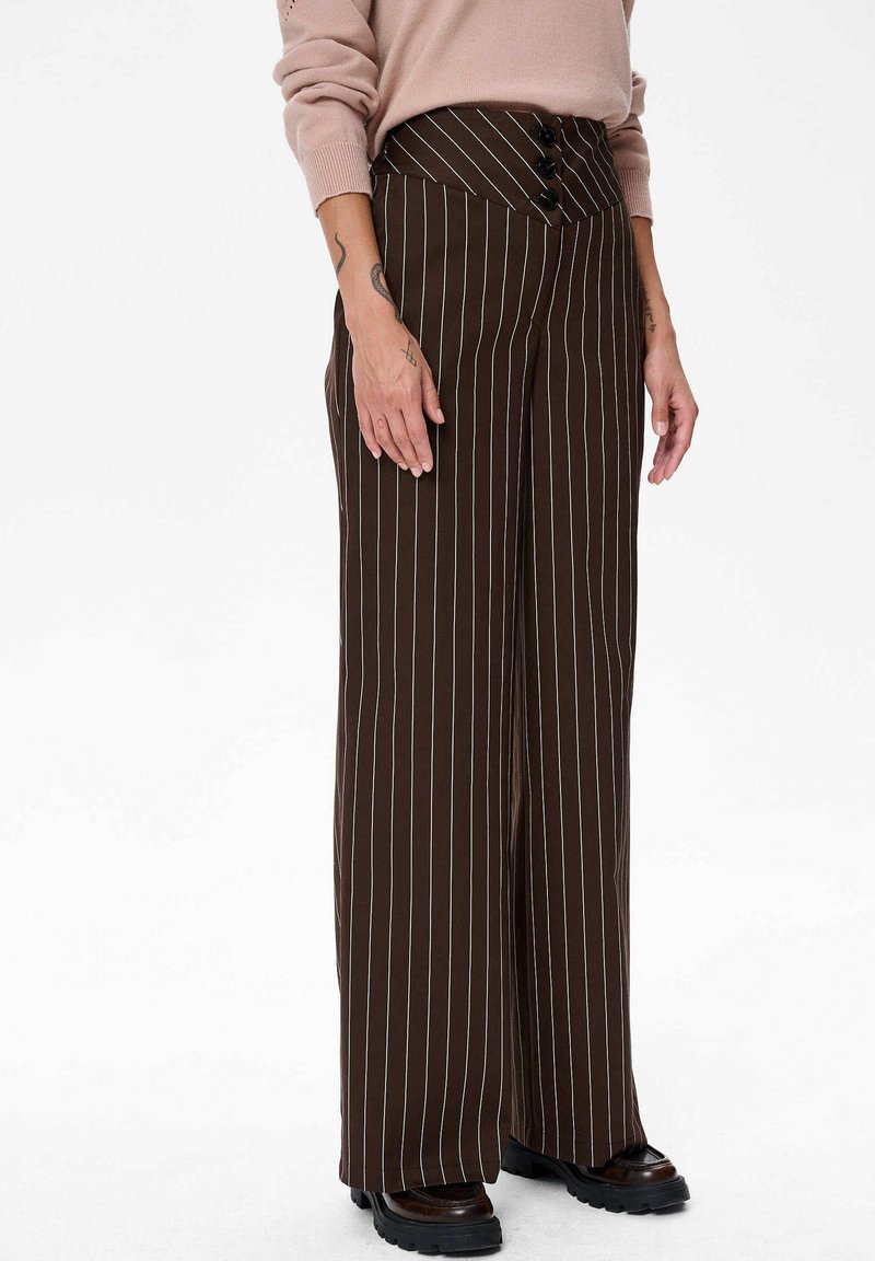 Vrouw in bruine hoge taille wijde broek met witte streepjes, drie zwarte knopen op de tailleband en zwarte chunky schoenen.