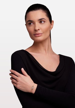 Un bracciale d'oro con un accento ovale colorato è indossato su una mano poggiata su una maglietta nera a maniche lunghe, drappeggiata, su uno sfondo chiaro.