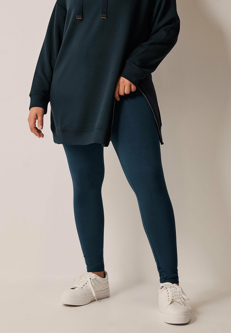 Sudadera oversized en verde azulado oscuro con puños acanalados, combinada con leggings a juego. Zapatillas blancas completan el look, con una suela texturizada.
