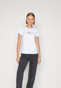 Biely bavlnený tričko s šedým logom 'ck' na prednej strane, v kombinácii s tmavošedými rovnými džínmi. Jednoduchý dizajn, krátke rukávy.