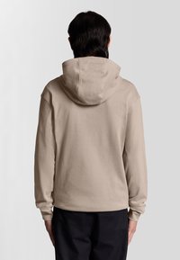 Beige hoodie med dragsko i huvan, mjuk textur, långa ärmar, avslappnad passform och ribbade muddar. Inga synliga logotyper eller mönster.
