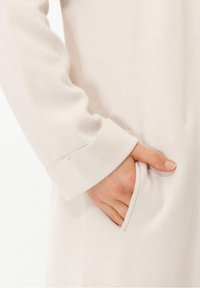 Jasno beżowy sweter z teksturowanej tkaniny, z podwiniętym mankietem i boczną kieszenią, o gładkim wykończeniu i miękkim wyglądzie.