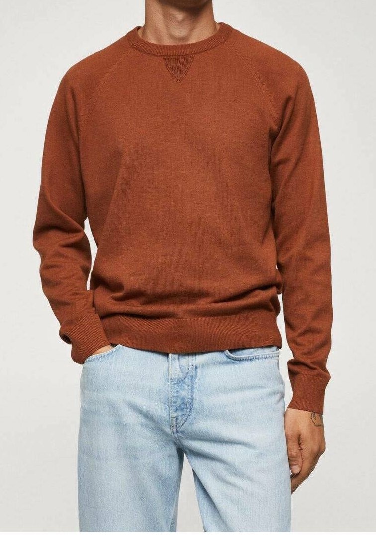 Homme portant un pull à manches longues couleur rouille et un jean bleu clair, une main dans la poche et un petit tatouage sur le poignet.