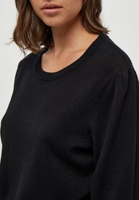 Maglione nero a maglia con colletto rotondo, maniche leggermente a sbuffo e una texture liscia, con una silhouette aderente.