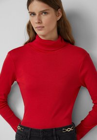 Frau mit langen, braunen Haaren, die einen roten Rollkragenpullover und schwarze Hosen mit goldener Kette am Hosenbund trägt.