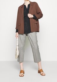 Person klädd i brun kavaj, svart blus, grå rutiga cropped byxor, bruna klacksandaler och håller en vävd krämfärgad handväska.