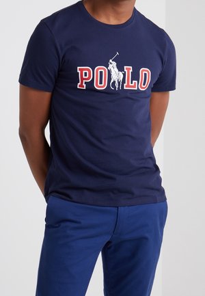 Mężczyzna ubrany w granatową koszulkę z czerwonym i białym napisem "POLO" oraz grafiką jeźdźca, do której dobiera niebieskie spodnie.