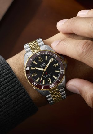 Persona pielāgo Timex Deepwater pulksteni ar melnu ciparnīcu, zelta un sudraba metāla siksnu un ūdensizturību līdz 200 m uz savas rokas.