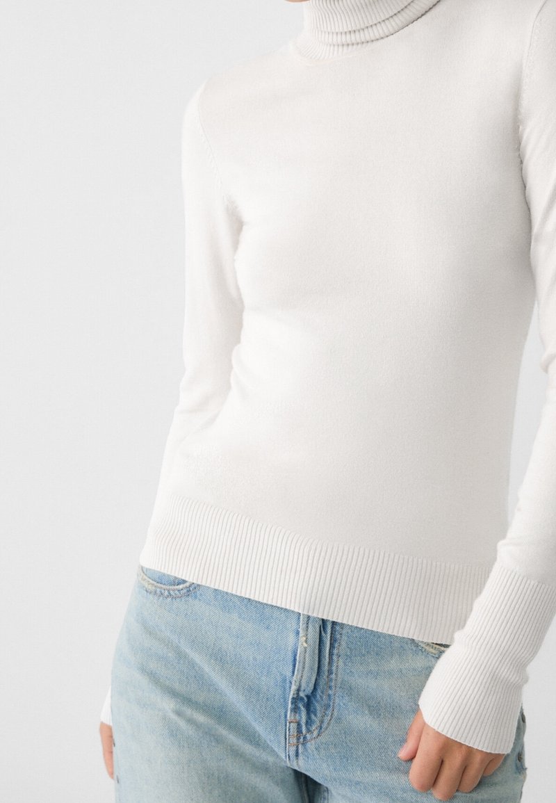 Personne portant un pull à col roulé côtelé blanc et un jean bleu clair, une main détendue le long du corps, sur un fond uni.