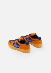 Baskets en suède orange et marine avec des accents violets, langue en maille texturée, semelles en caoutchouc, détails perforés et lacets contrastés.