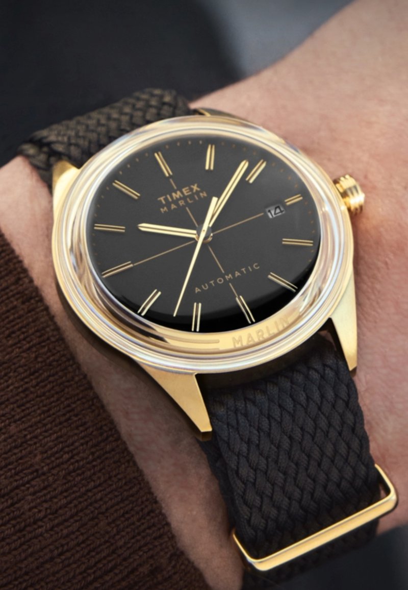 Automatski sat Timex Marlin s crnim brojčanikom, zlatnim markerima i teksturiranom tamnom tkanom narukvicom. Ima prozor za prikaz datuma na 3 sata.
