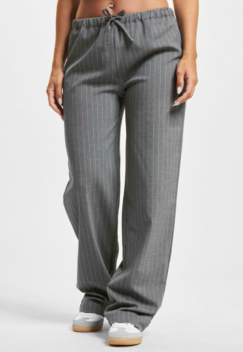 Femme portant un pantalon ample gris à fines rayures avec taille à cordon et baskets blanches, debout devant un fond uni.