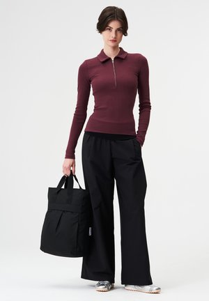 Frau trägt ein burgunderrotes langärmliges Oberteil und schwarze Hosen mit weitem Bein, hält eine schwarze Tragetasche und steht vor einem weißen Hintergrund.