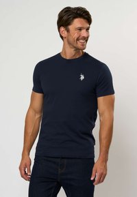 Tamsiai mėlynas medvilninis marškinėlis su apvalia iškirpte ir trumpomis rankovėmis, ant kairio krūtinės mažas baltas logotipas. Lygus audinys.