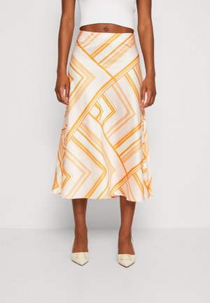 Vrouw draagt een witte en oranje midi-rok met geometrisch patroon, een wit cropped mouwloos topje en crèmekleurige puntige pumps.