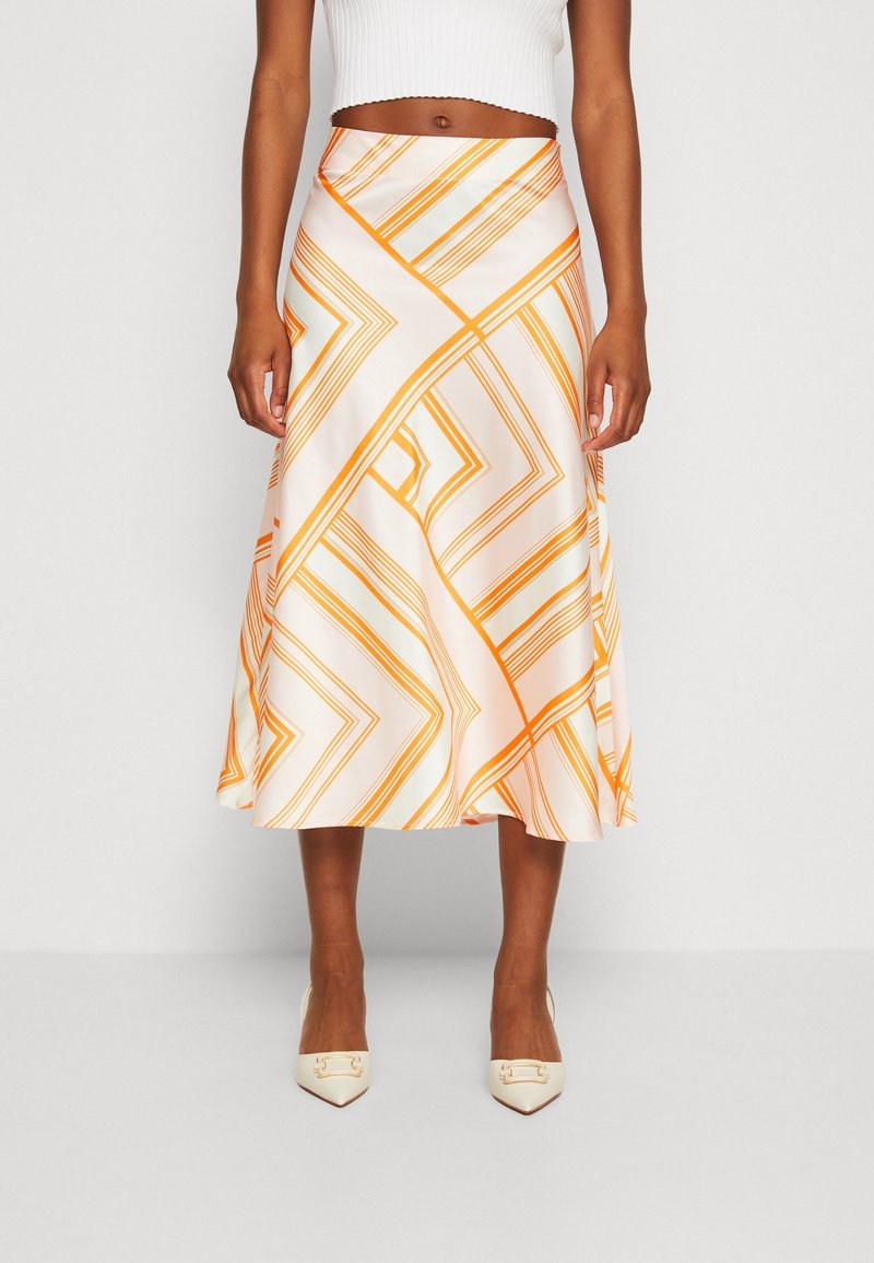 Femme portant une jupe midi à motif géométrique blanc et orange, un top court sans manches blanc et des escarpins à bout pointu couleur crème.
