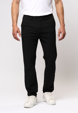 Man draagt zwarte slim-fit broek met riemlussen, witte sneakers en een wit T-shirt, staand tegen een effen achtergrond.