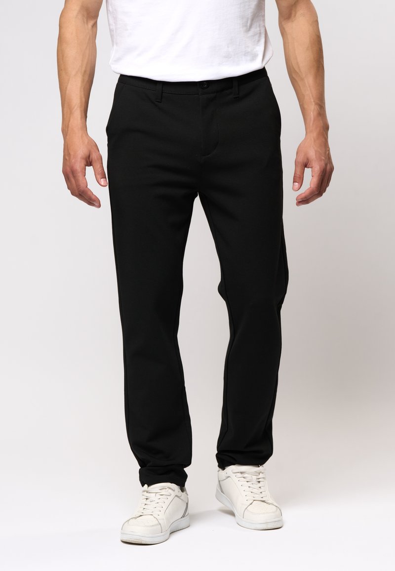 Homme portant un pantalon noir slim avec passants pour ceinture, des baskets blanches et un t-shirt blanc, debout devant un fond uni.