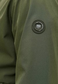 Maslinasto zelena jakna s glatkom teksturom i zaobljenim zakrpom s natpisom "JACK & JONES" na gornjem rukavu.