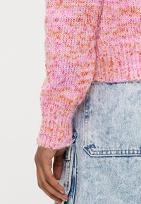 Adīts džemperis rozā krāsā ar oranžiem plankumiem, ar teksturētu rakstu un ribveida aprocēm. Kombinēts ar gaiši zilu džinsa apģērbu.