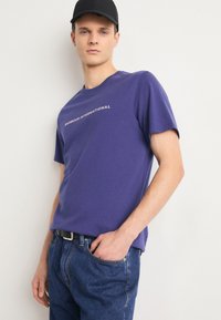 Purple cotton t-shirt features the text "BARBOUR INTERNATIONAL" in white across the chest. Paired with blue denim jeans and a black cap.