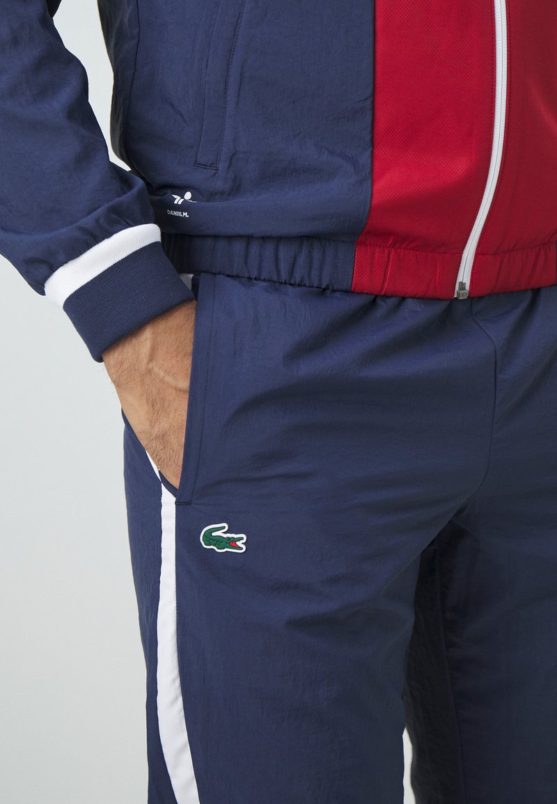 Survêtement bleu marine avec des accents rouges, comprenant une veste zippée et une ceinture élastique. Comprend un logo crocodile vert et blanc sur le pantalon.