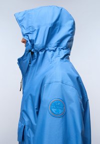 Person iført en blå hættetrøje med en rund lap på venstre ærme, der vender mod venstre mod en ensfarvet lys baggrund.