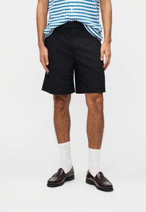 Uomo con pantaloncini neri fino al ginocchio, calzini bianchi alla caviglia, mocassini in pelle marrone scuro e una maglia a righe blu e bianche.