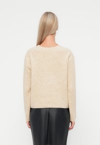 Bežs ribots adījuma džemperis ar apaļu kakla izgriezumu un garām piedurknēm, apvienots ar melnu ādas svārku. Redzama maiga tekstūra.