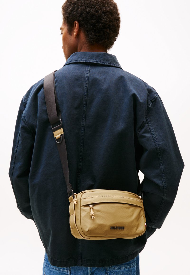 Borsa a tracolla beige con una texture liscia, chiusura con zip e tracolla regolabile. Marchiata con "HILFIGER" in nero, esposta su una giacca scura.
