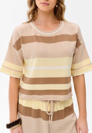 Kobieta nosząca dzianą beżową, brązową i żółtą koszulkę w paski z krótkim rękawem oraz pasujące szorty ze sznurkiem, z kolczykami i futrzaną bransoletką.