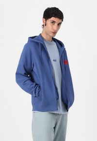 Blå zip-up hettegenser med frontlomme og snor i hetten, kombinert med en lyseblå T-skjorte. Har røde og blå logo-lapper.