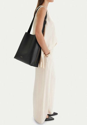 Mujer con top sin mangas beige y pantalones anchos, llevando un gran bolso tote negro de cuero con cremallera lateral y logo dorado al hombro.