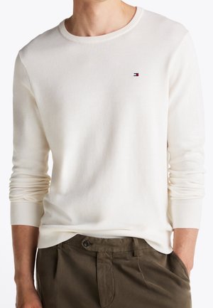 Homme portant un pull blanc uni à manches longues avec un petit logo sur la poitrine et un pantalon vert olive, mains dans les poches.