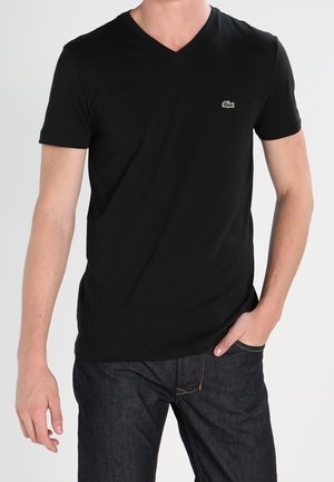 Hombre vistiendo una camiseta negra de cuello en V con un pequeño logo de cocodrilo verde, jeans azul oscuro, una mano en el bolsillo, contra un fondo claro y simple.