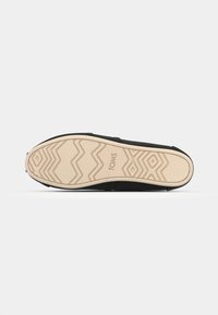 Sorte TOMS-såle med en teksturert gummioverflate med sikksakkmønster og en kremfarget bunn. Logoen "TOMS" er synlig.