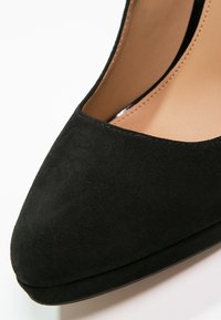 Chaussure à talon haut en suède noir avec bout pointu et doublure intérieure beige, présentée en gros plan sur un fond blanc.