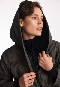 Pikowany zielony kurtka puchowa z dużym kapturem i czarnym futrem sztucznym. Posiada haftowane detale na powierzchni materiału.