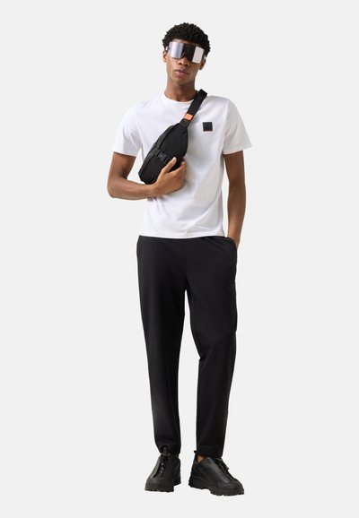 Homme portant un t-shirt blanc, un pantalon noir, des baskets noires, de grandes lunettes de soleil réfléchissantes et un sac bandoulière noir, debout devant un fond uni.