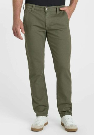 Mann trägt olivgrüne gerade Hose und weiße Sneakers, steht mit einer Hand in der Tasche vor einem schlichten Hintergrund.