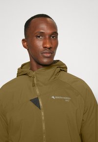 Khaki hooded jacket featuring a zip closure, textured black accents, and a logo. The fabric appears lightweight and weather-resistant.