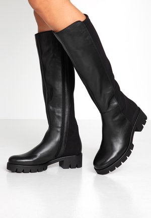Bottes en cuir noires montantes jusqu'aux genoux avec semelles épaisses et fermetures éclair latérales, portées sur des jambes nues sur un fond blanc.